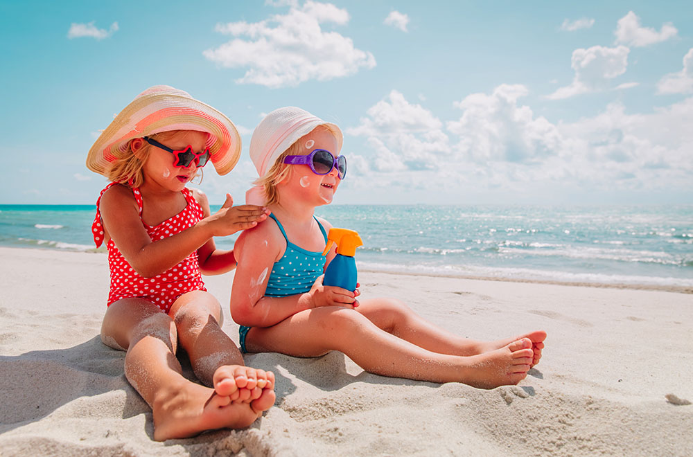 Dve dekleti na plaži v kopalkah, z zaščitnimi očali in klobuki, ki se nanašata s kremo za sončenje.