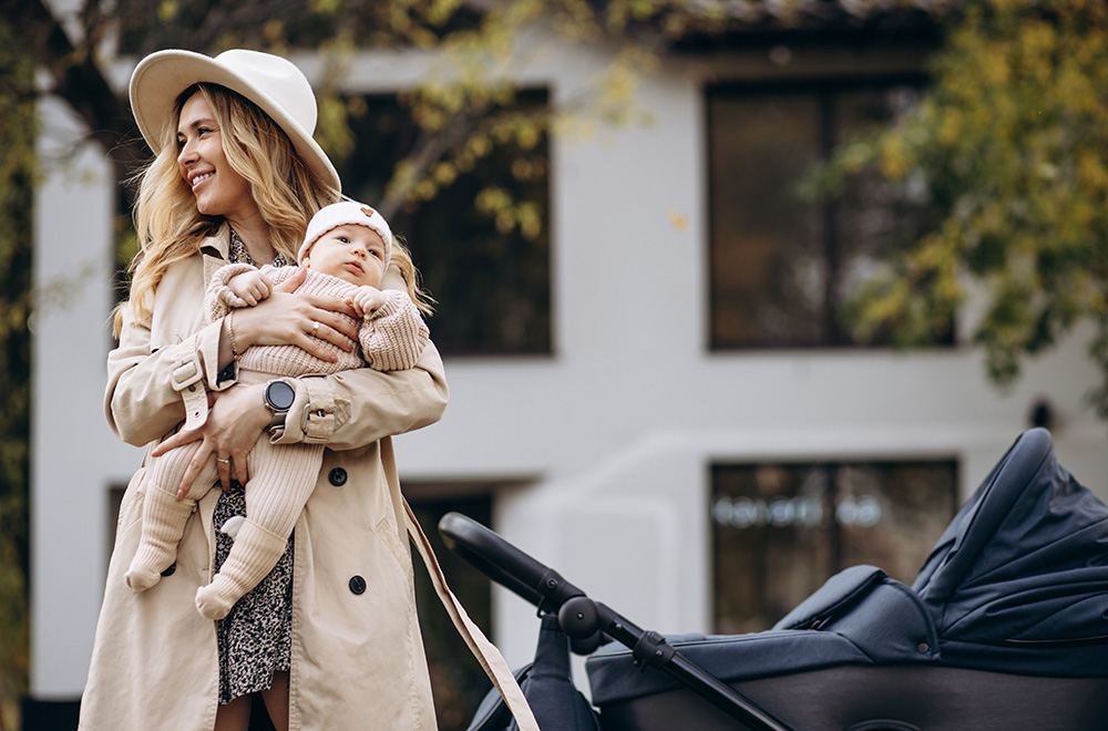 Ženska v naročju drži dojenčka in hodi ob vozičku pred stavbo.